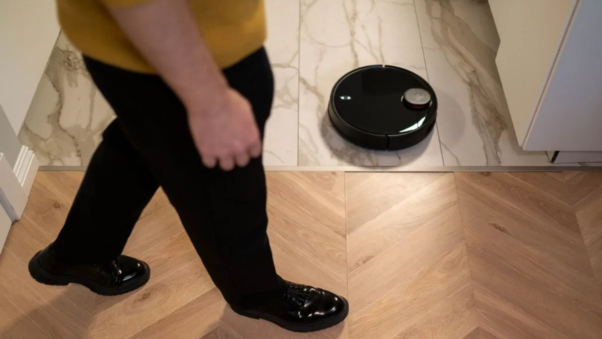 Pessoa caminha no corredor enquanto um robô aspirador preto limpa o piso de mármore em uma casa moderna.