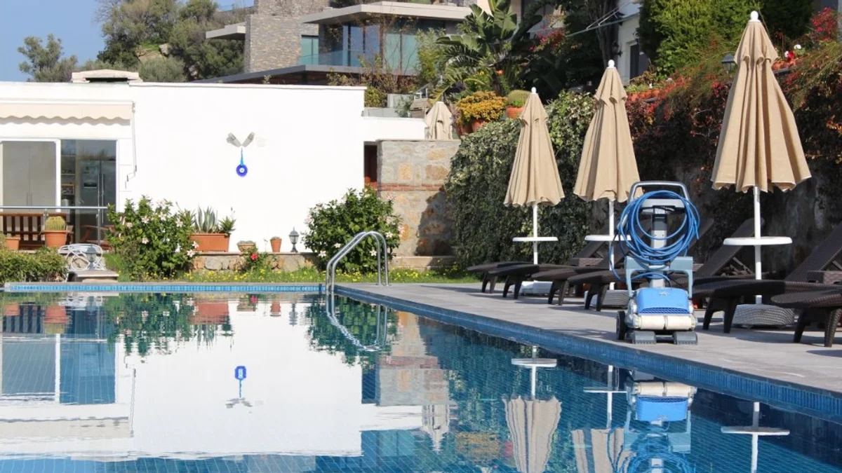 Robô aspirador de piscina azul e branco com cabo enrolado sobre deck de pedra ao lado da piscina com água azul e reflexos.