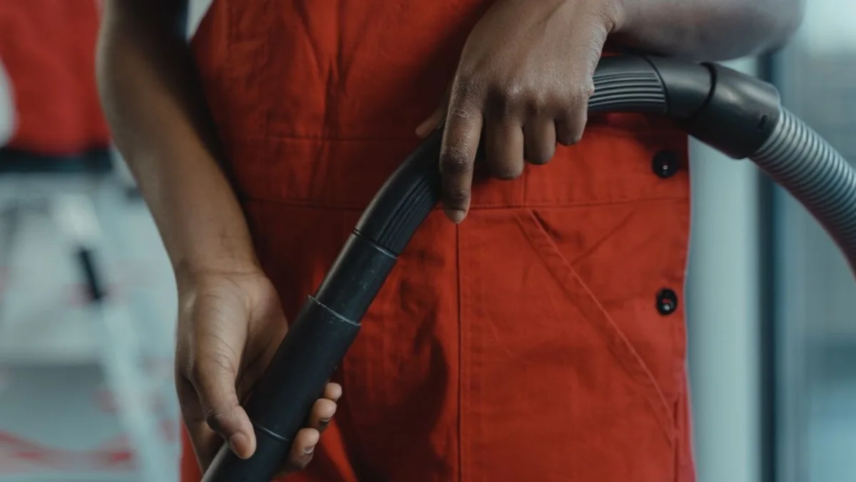Mãos seguram a mangueira flexível preta de um aspirador de pó durante a limpeza, com macacão vermelho visível no enquadramento e ambiente interno ao fundo.