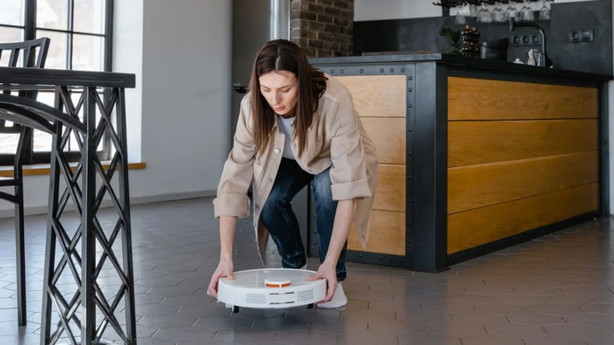 Mulher agachada em cozinha moderna segurando robô aspirador branco com sensor laranja sobre piso de ladrilhos cinzas hexagonais.