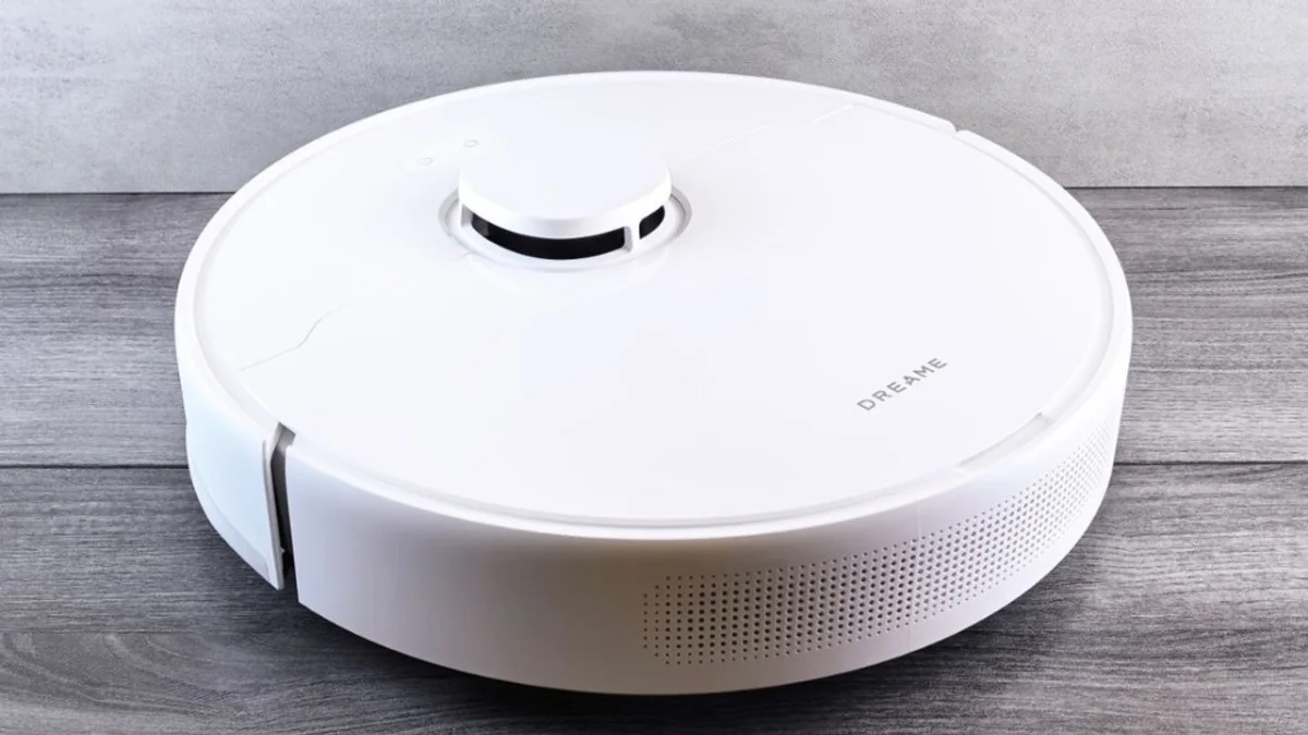 Robô aspirador circular branco com torre de sensor LiDAR sobre piso de madeira escura, em ambiente interno com parede cinza.