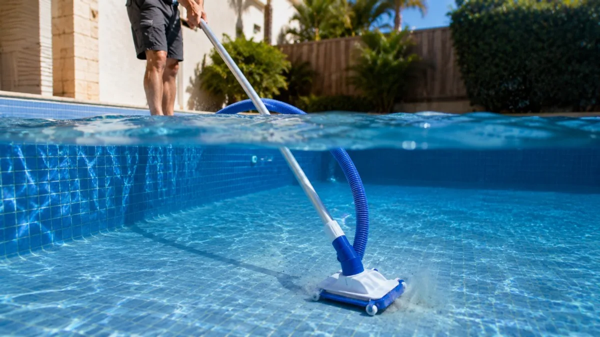 Aspirador manual genérico deslizando lentamente pelo fundo de piscina residencial com água azul cristalina, preso ao cabo telescópico.